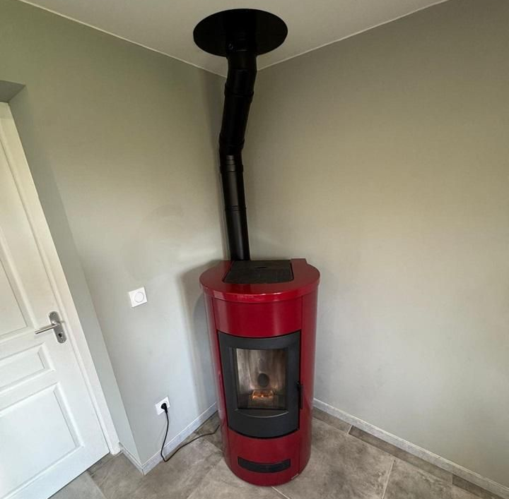 Poêle rouge à conduit de cheminée noir, placé dans un coin aux murs et au sol gris.