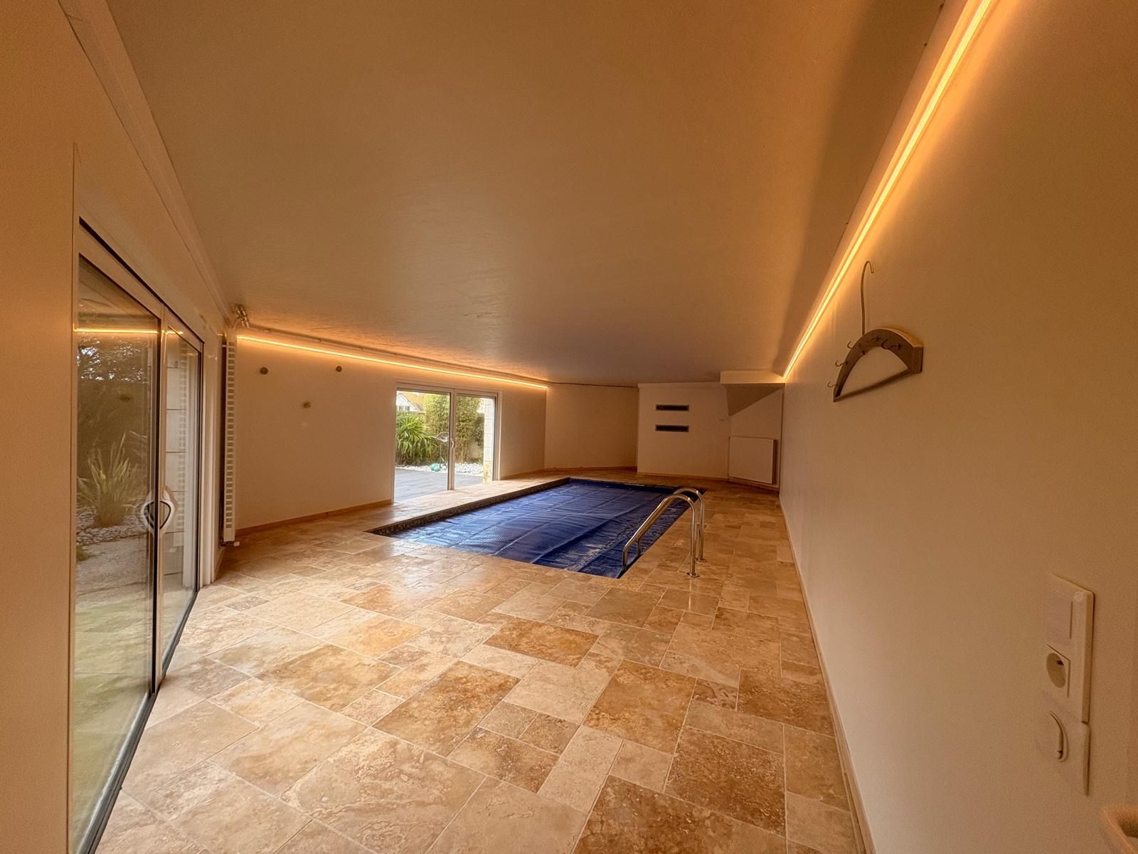 Salle de piscine intérieure avec sol en travertin, grandes fenêtres et couverture de piscine bleue.