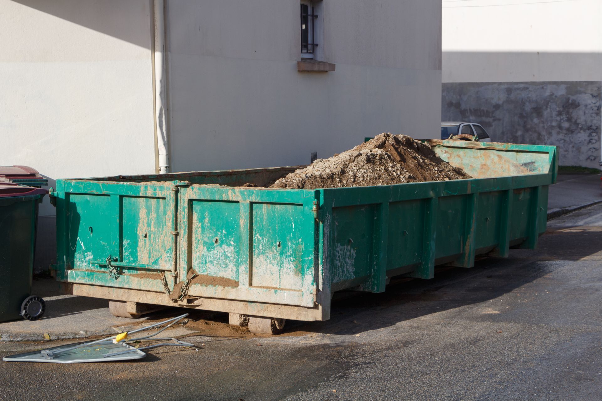 Un conteneur à déchets de chantier vert, rempli de terre, est posé sur une surface pavée près d'un bâtiment blanc.