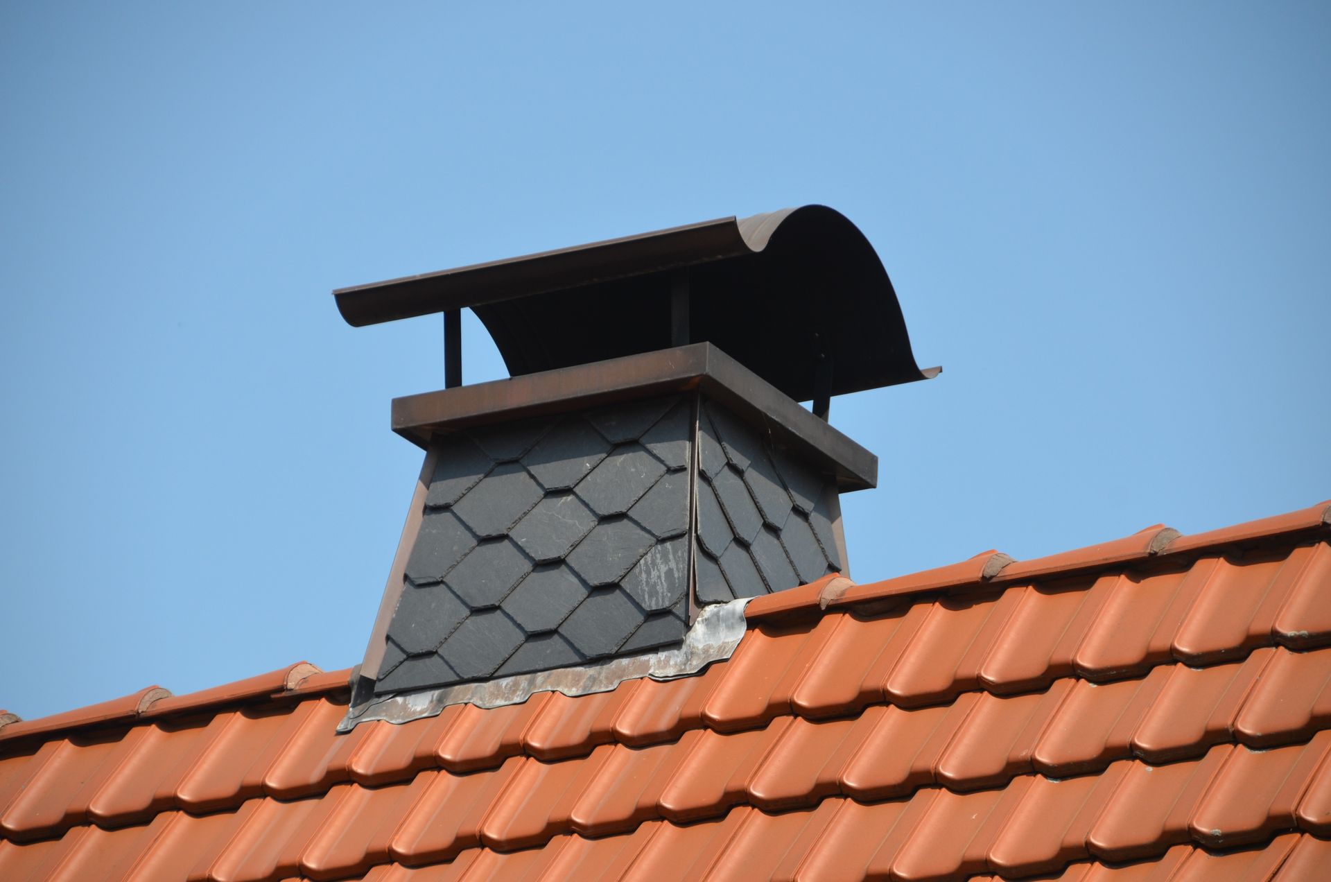 Cheminée sur un toit de tuiles rouges, recouvert d'ardoises sombres, avec un chapeau de pluie métallique incurvé.