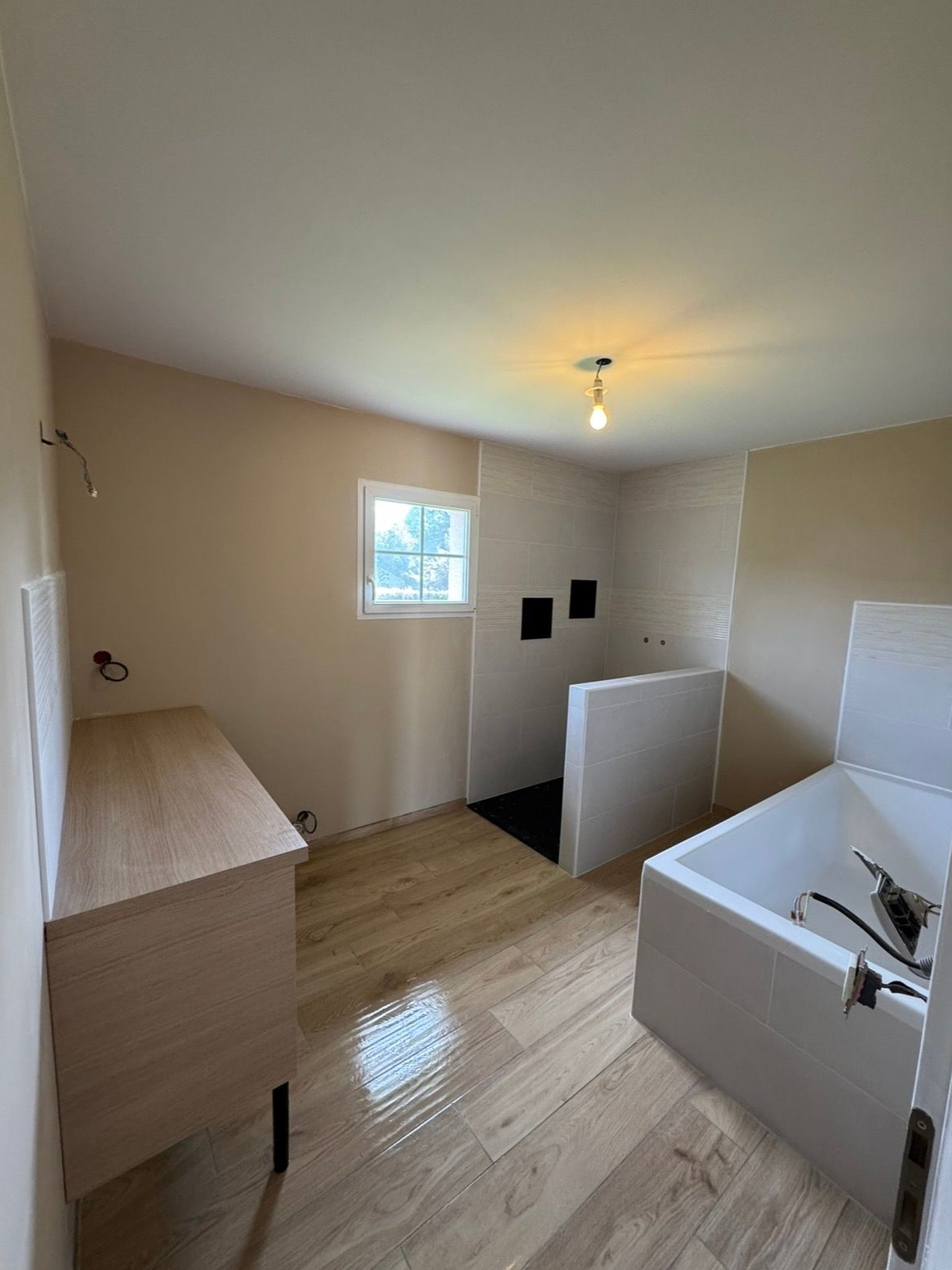 Pièce beige vide avec parquet, petite fenêtre, plafonnier et éléments ou cloisons blancs.