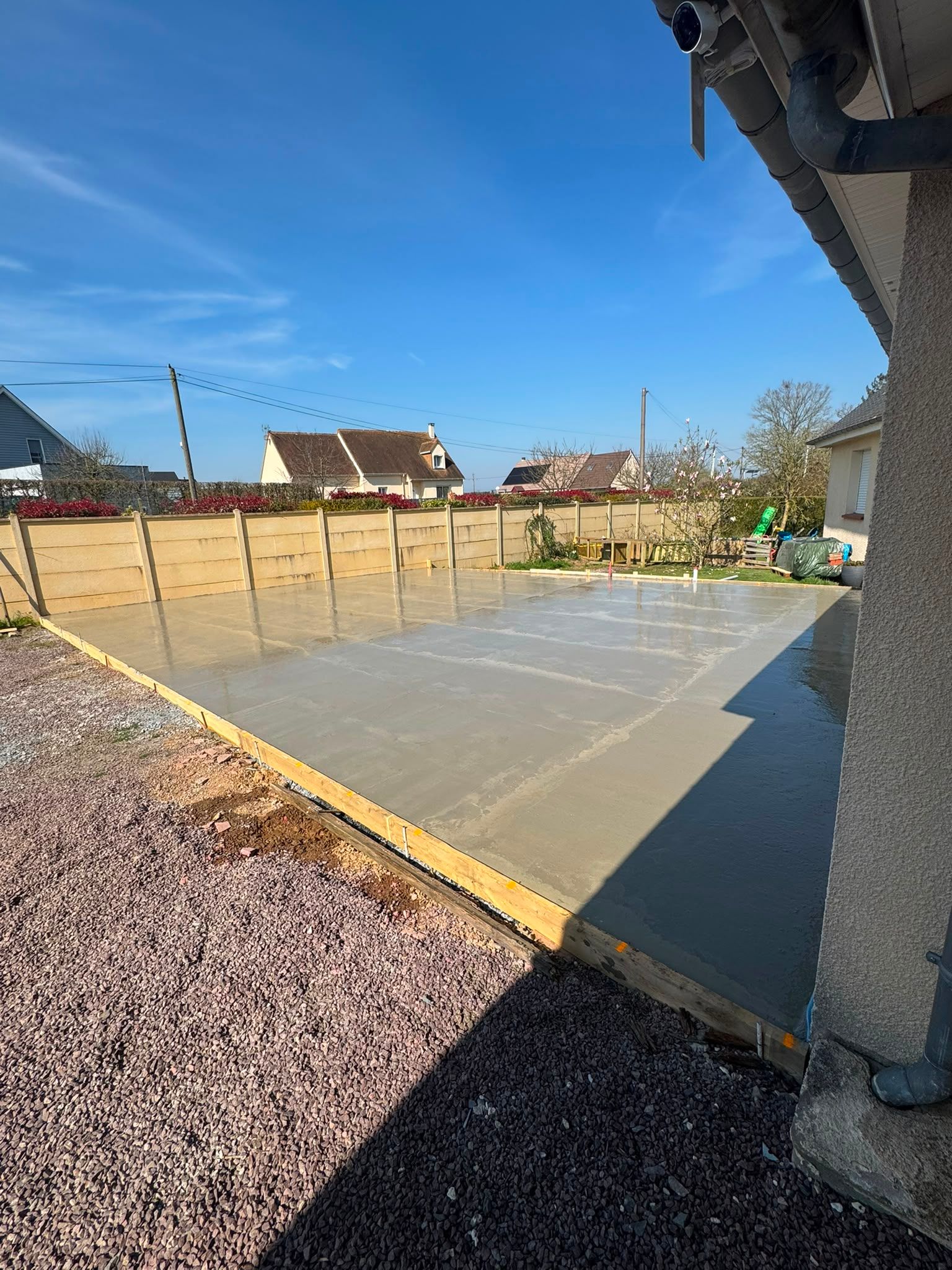 Dalle de béton fraîche dans une cour arrière à côté d'une maison, bordée d'une clôture en bois sous un ciel bleu clair
