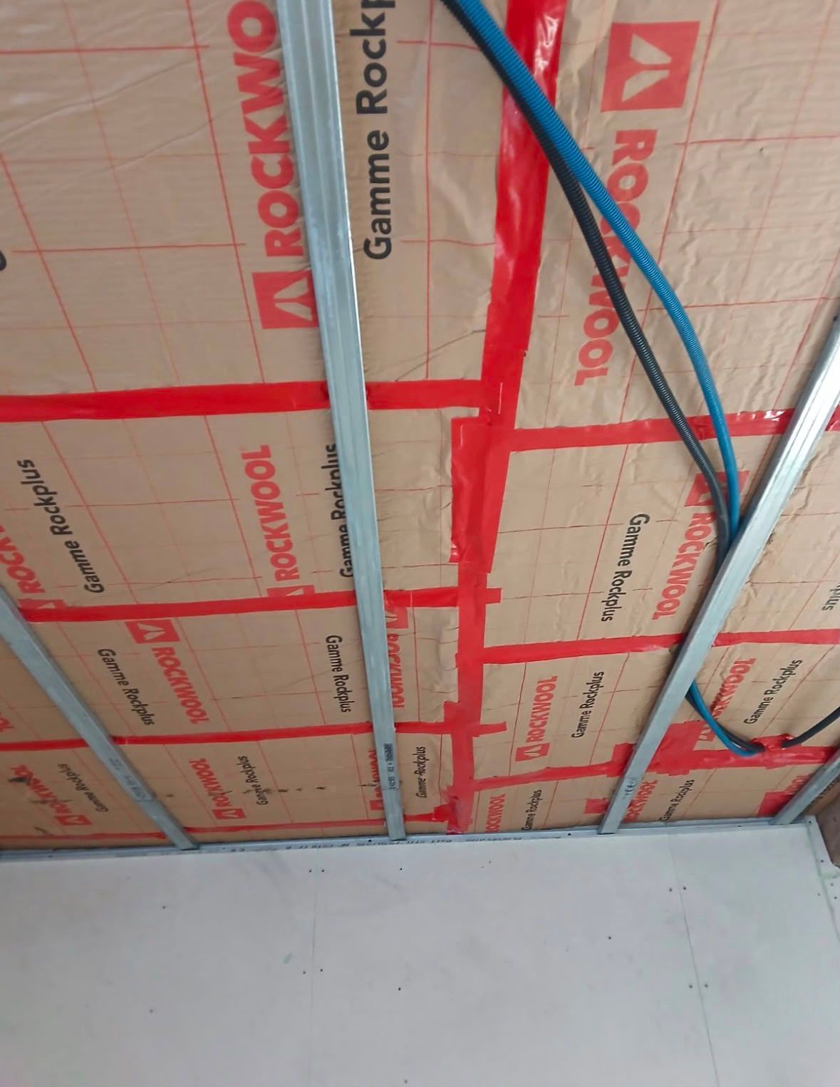 Plafond en construction avec ossature rouge, fils électriques bleus et blancs et panneaux isolants.
