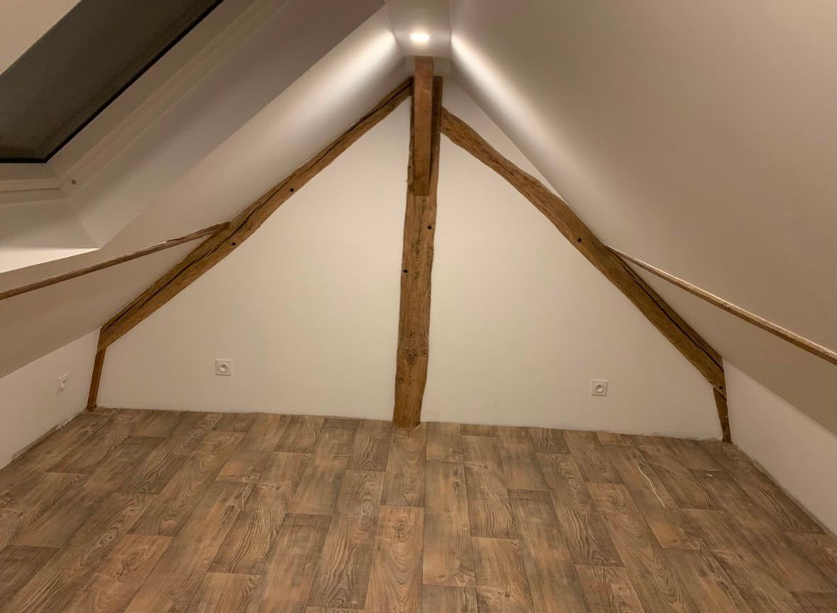 Petite pièce mansardée avec murs blancs en pente, poutres apparentes et parquet à motifs.