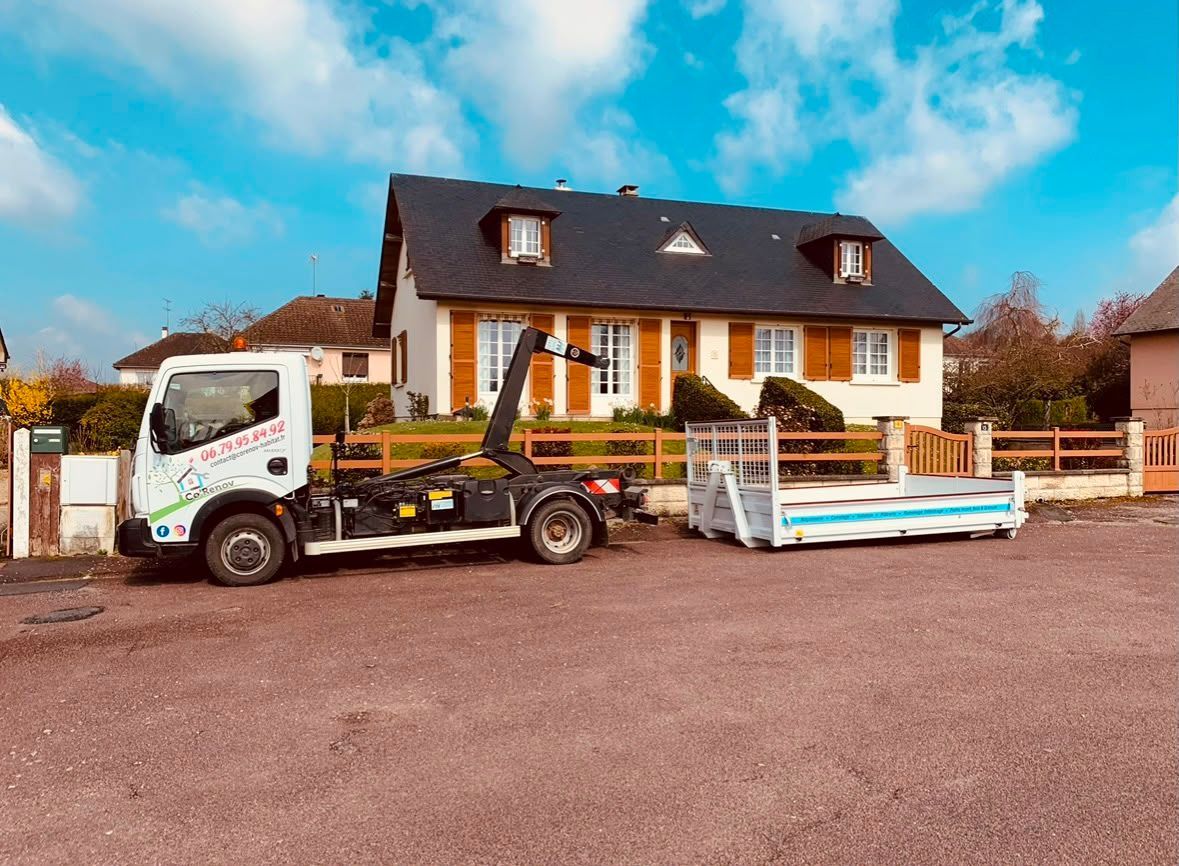 Un camion blanc tractant une remorque à plateau avec un bateau bleu devant une maison sous un ciel nuageux