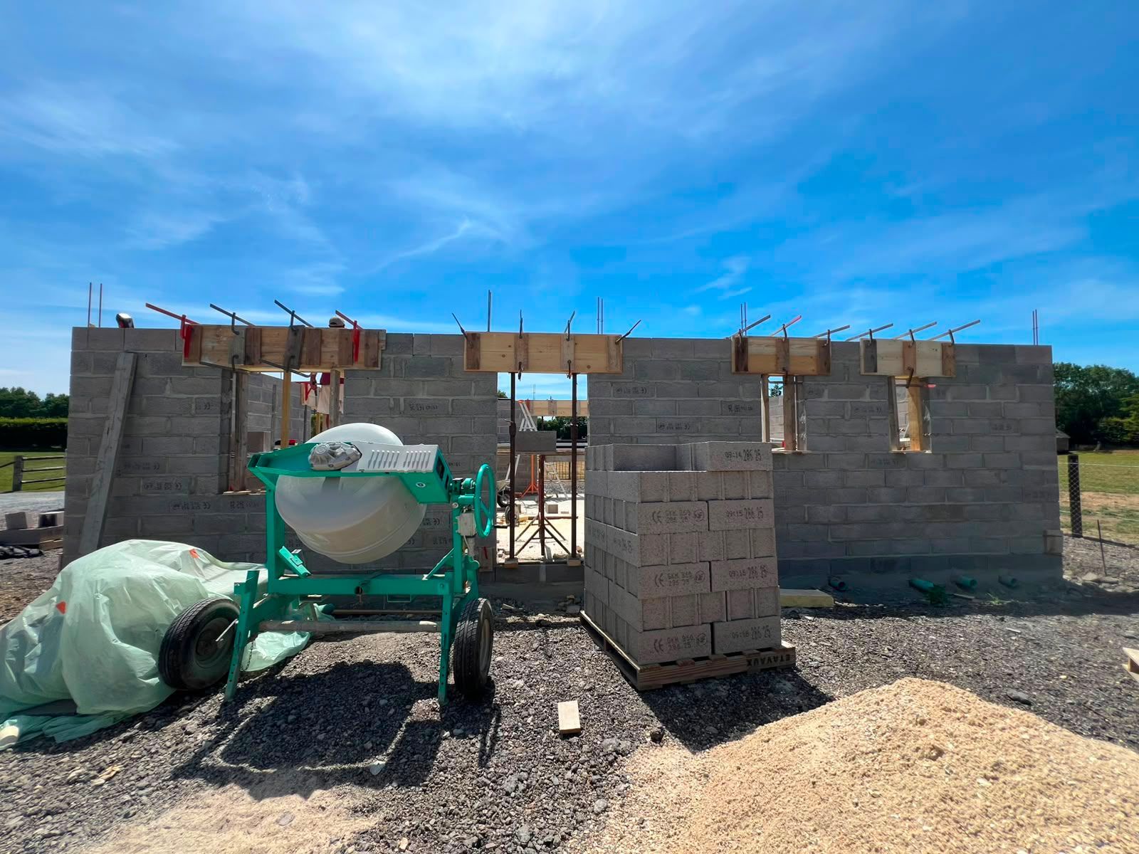 Chantier de construction avec une maison en parpaings partiellement construite, une bétonnière et des matériaux sous un ciel d'un bleu éclatant