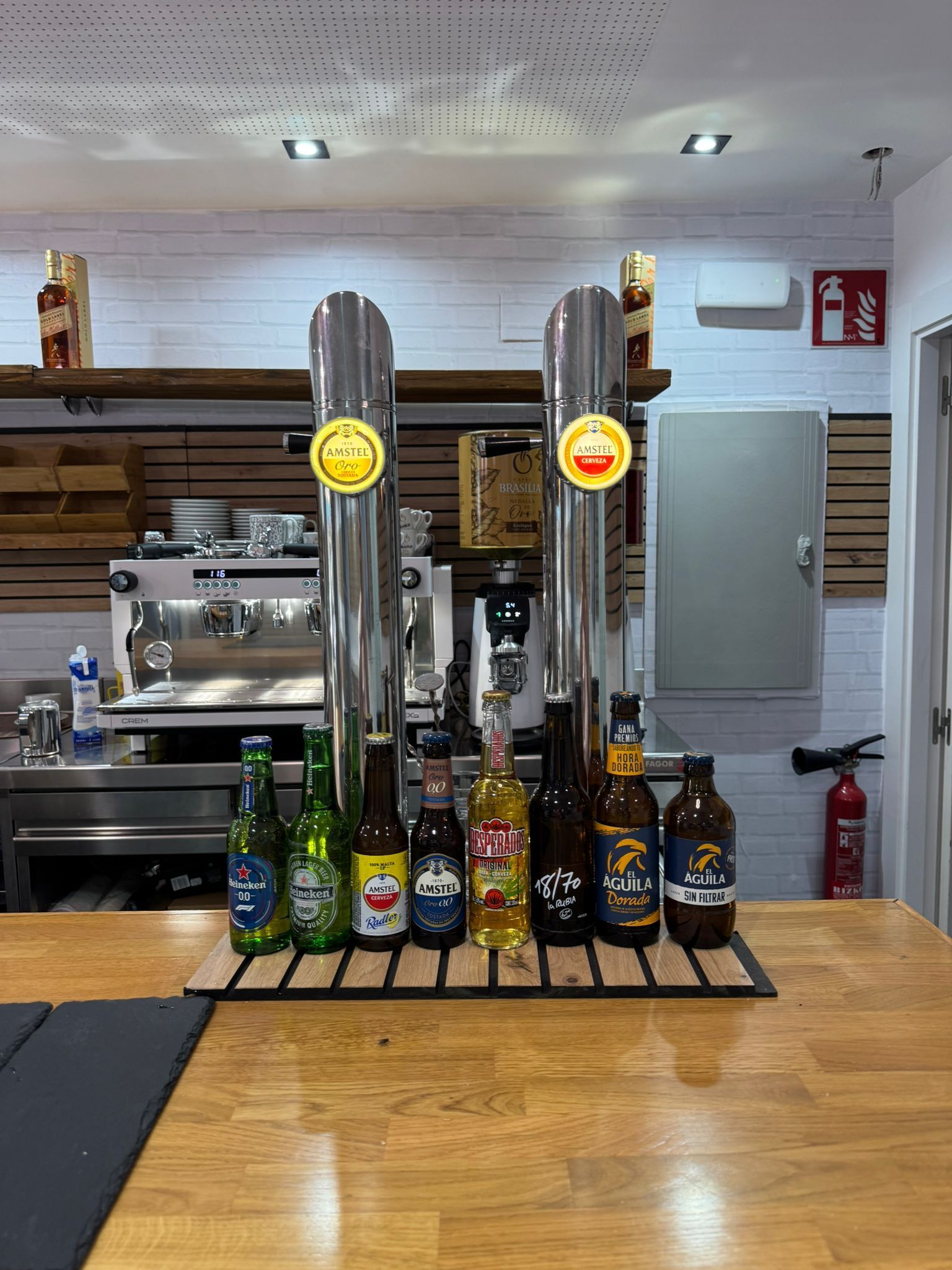 Un mostrador de bar con grifos de cerveza y diversas bebidas embotelladas, incluidas cerveza y licor, con un fondo de equipamiento de cocina.