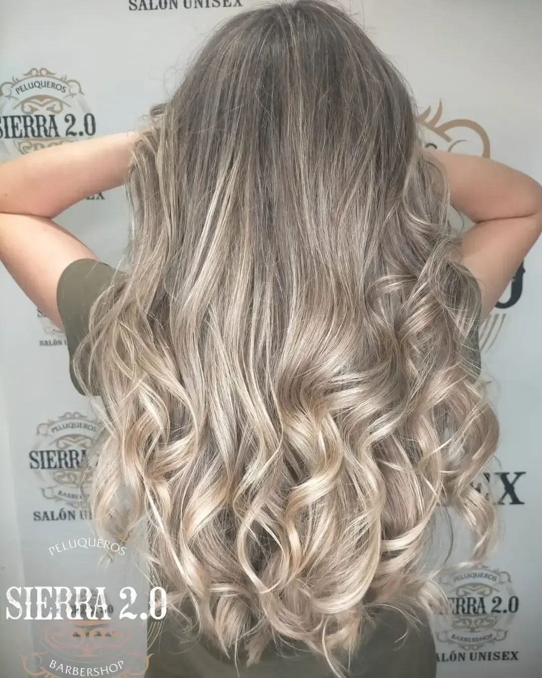 Peinado largo rubio ondulado con balayage visto desde atrás contra un fondo de salón.