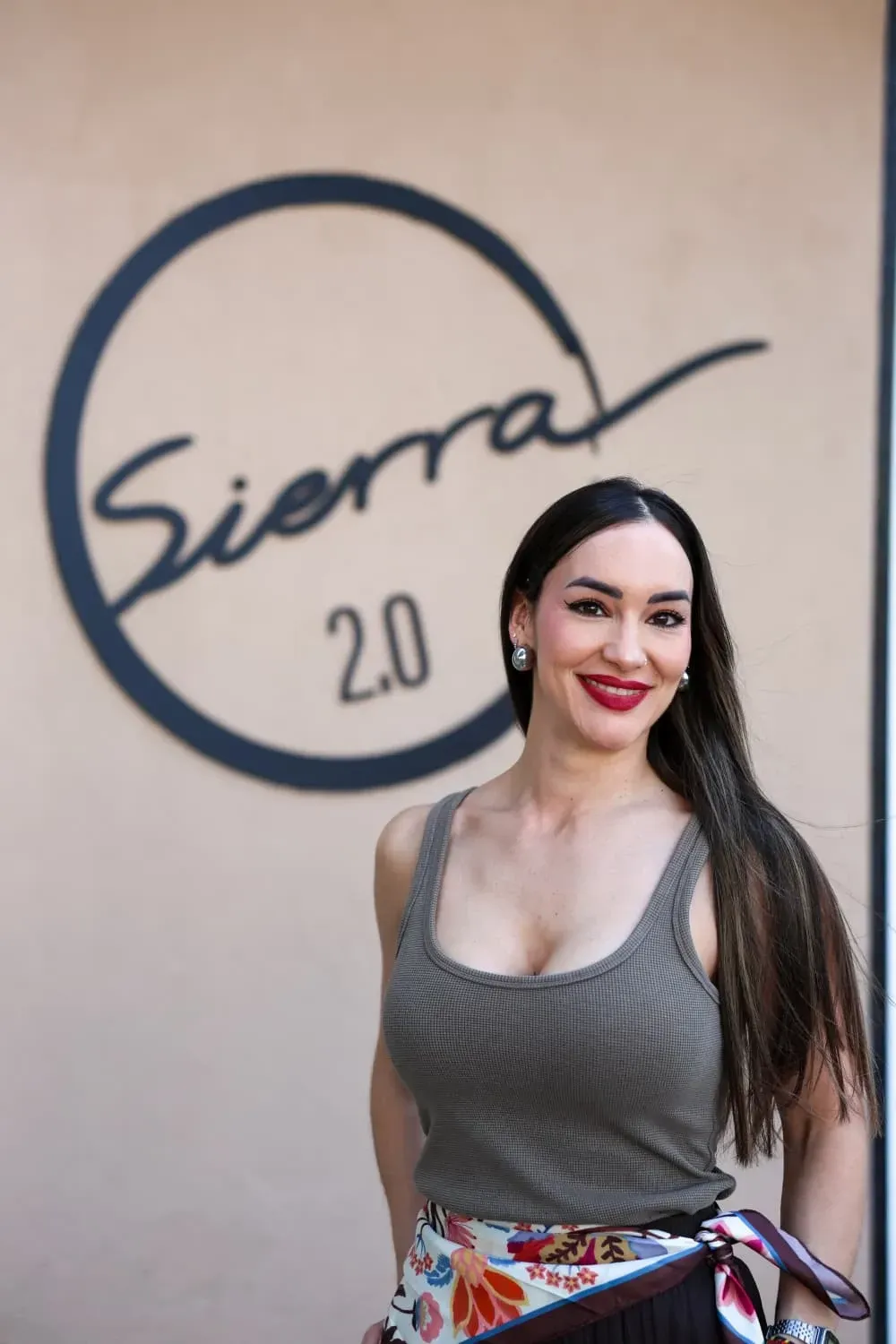 Mujer posando frente a un letrero de Sierra 2.0, con una blusa gris y una falda colorida.