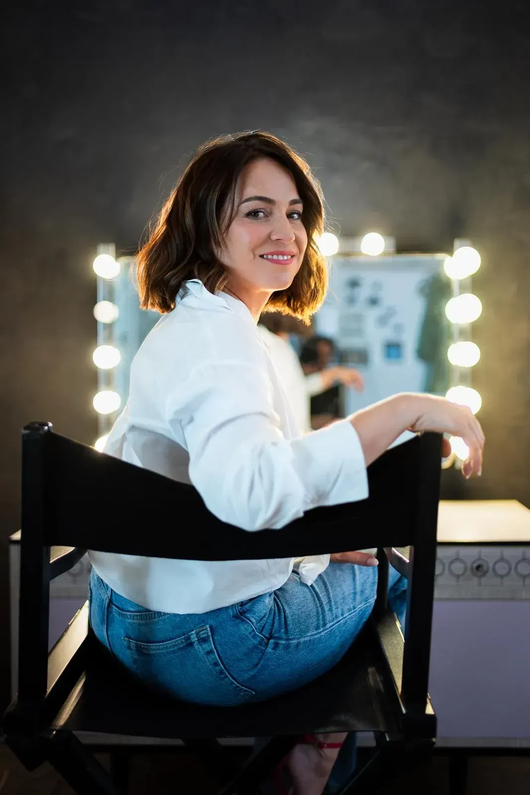 Una mujer sonríe sentada en una silla en un vestidor, con un espejo de tocador iluminado detrás de ella.