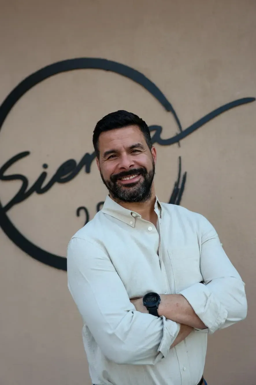 Hombre con camisa blanca sonriendo con los brazos cruzados frente a una pared con el logotipo de una cafetería.