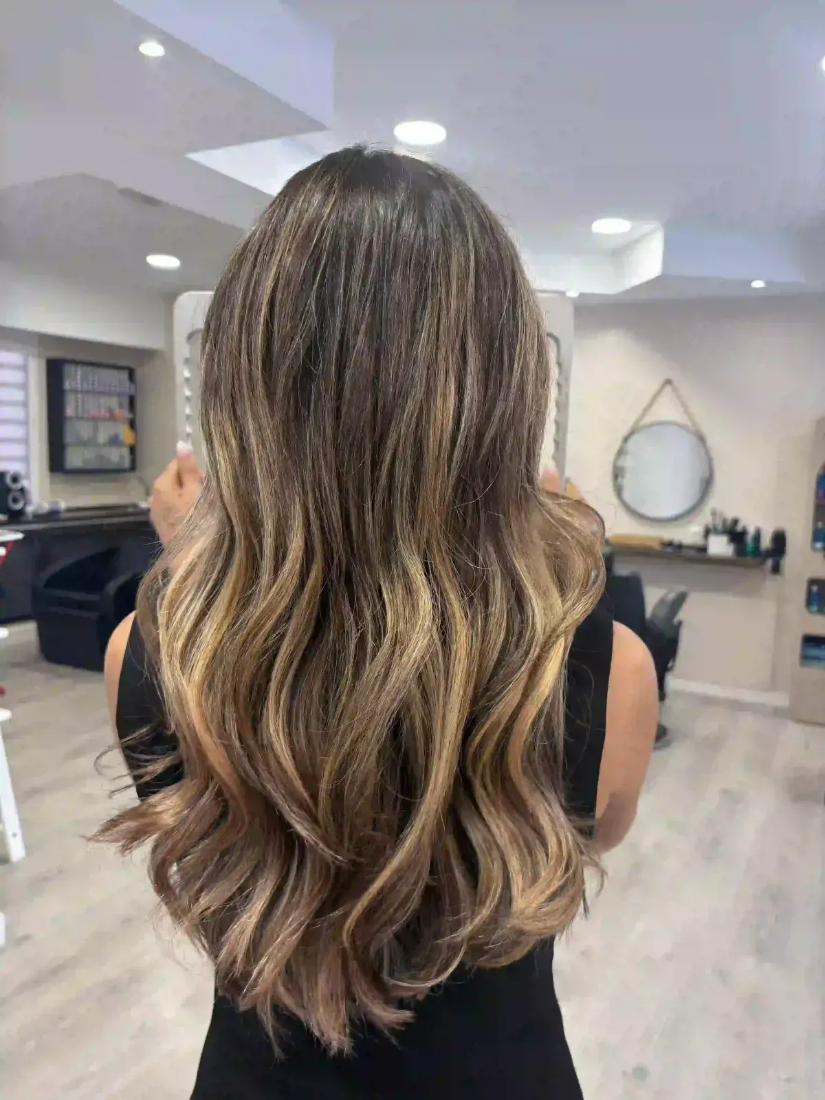 Cabello largo y ondulado con mechas balayage castaño visto desde atrás en el luminoso interior de un salón de belleza.