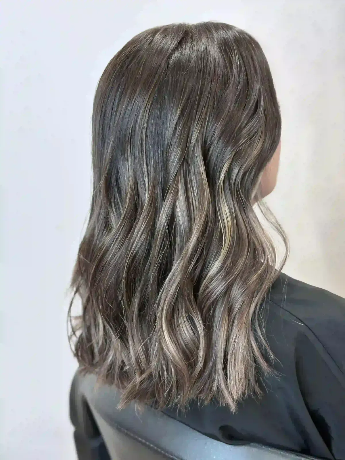 Vista posterior de cabello oscuro ondulado hasta los hombros con reflejos plateados sobre un fondo liso.