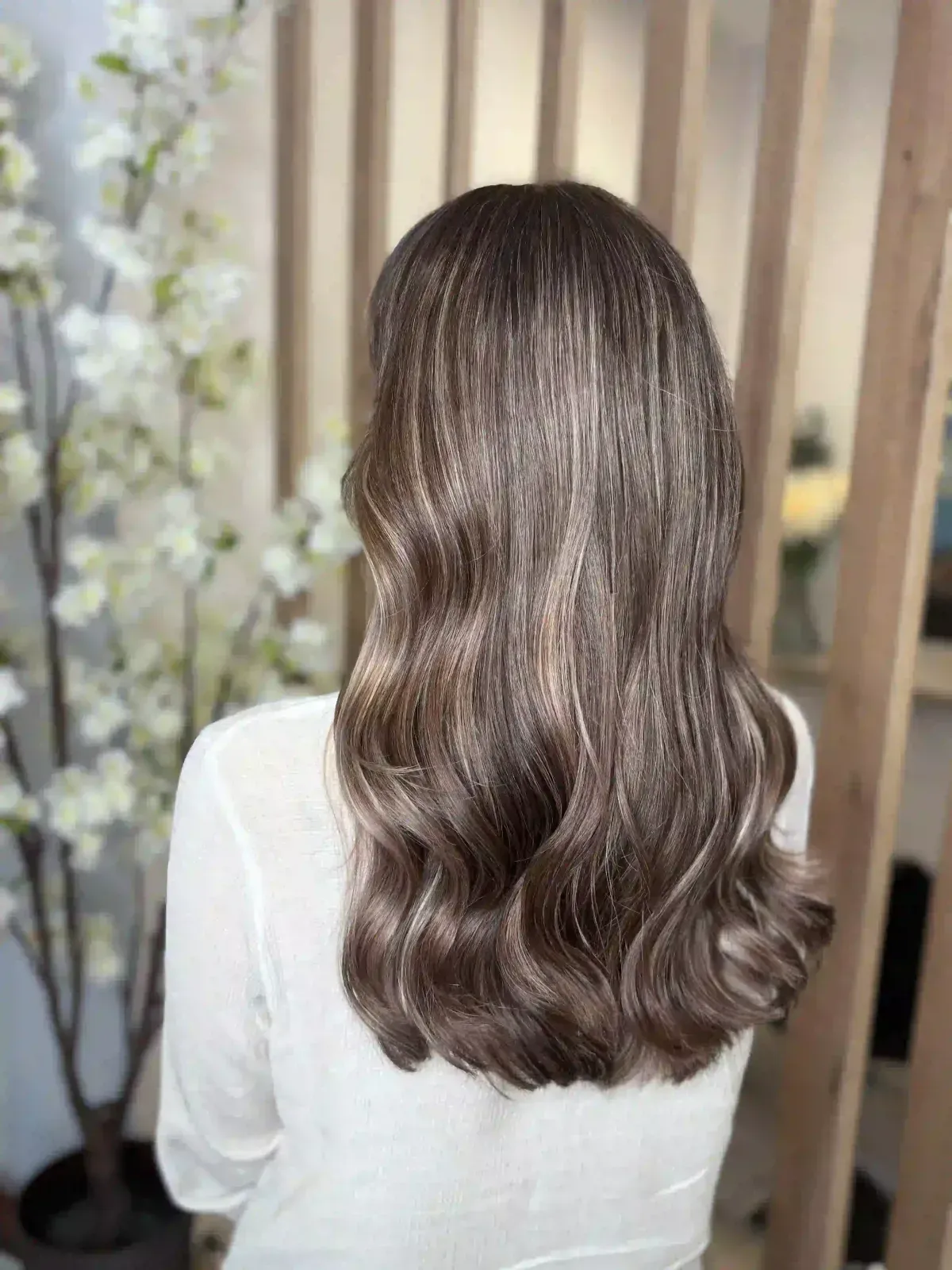 Cabello castaño largo y ondulado visto desde atrás, con una blusa blanca en un ambiente interior con iluminación tenue.