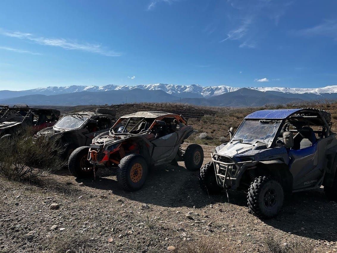 Cuatro vehículos todoterreno estacionados en un camino de tierra con montañas nevadas y un cielo azul de fondo.