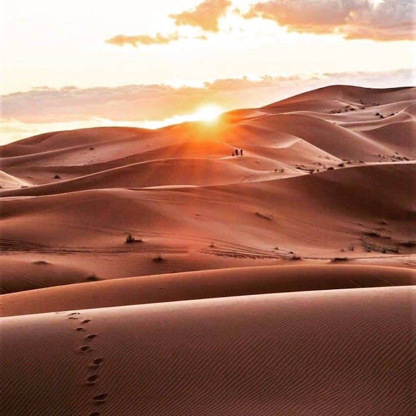 Atardecer sobre dunas de arena, con huellas en primer plano y un grupo de figuras recortadas en el horizonte.