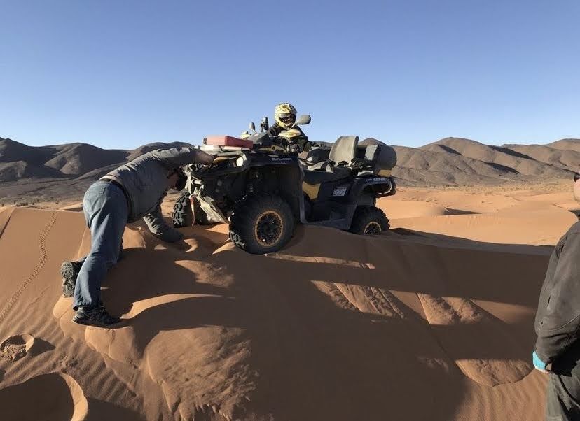 Un hombre empuja un quad atascado en una duna; otra persona está sentada sobre él. Escenario desértico, día soleado.