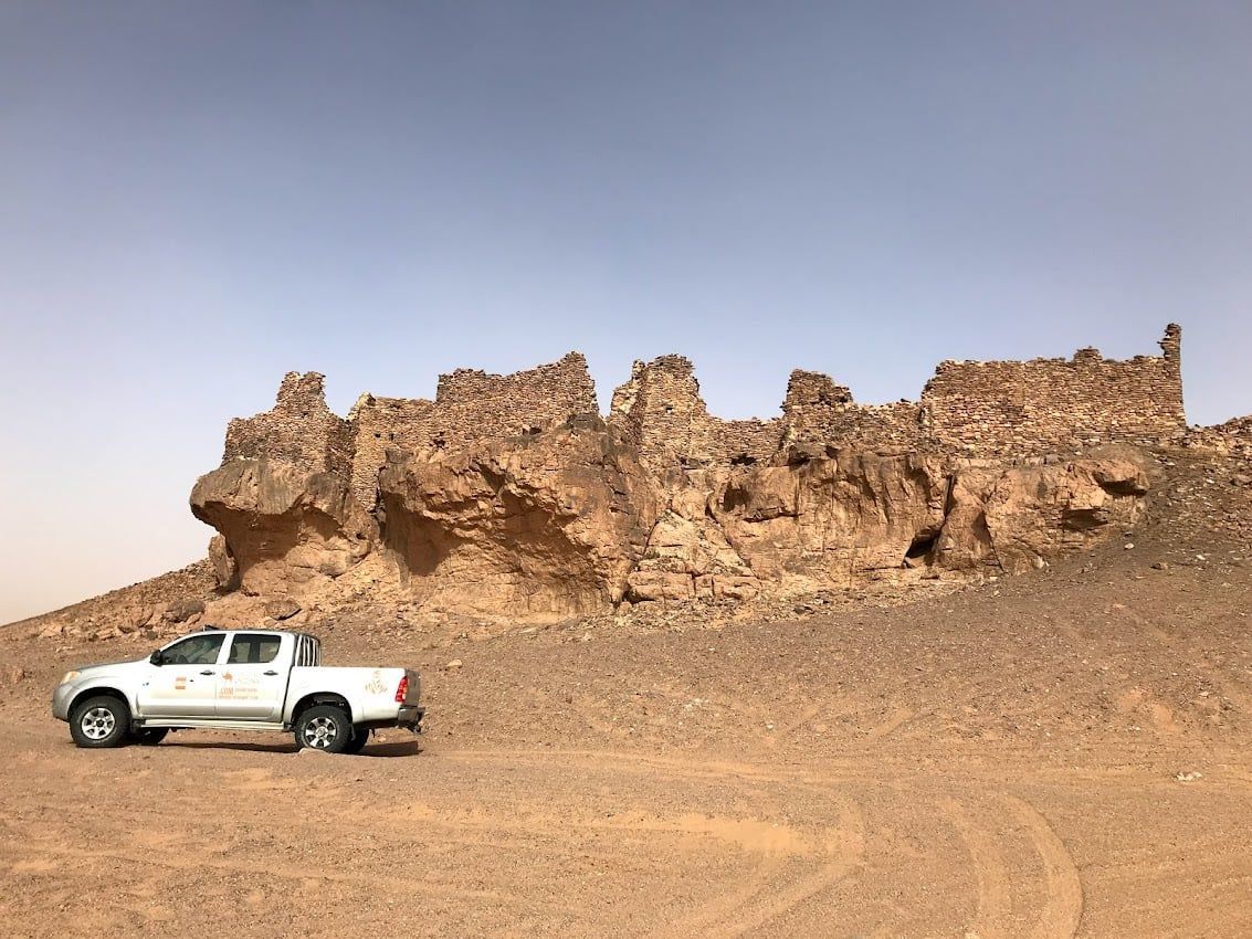 Camioneta blanca estacionada frente a antiguas formaciones rocosas desmoronadas en un paisaje desértico.