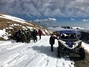 Personas y vehículos todoterreno en un sendero nevado de montaña bajo un cielo azul.