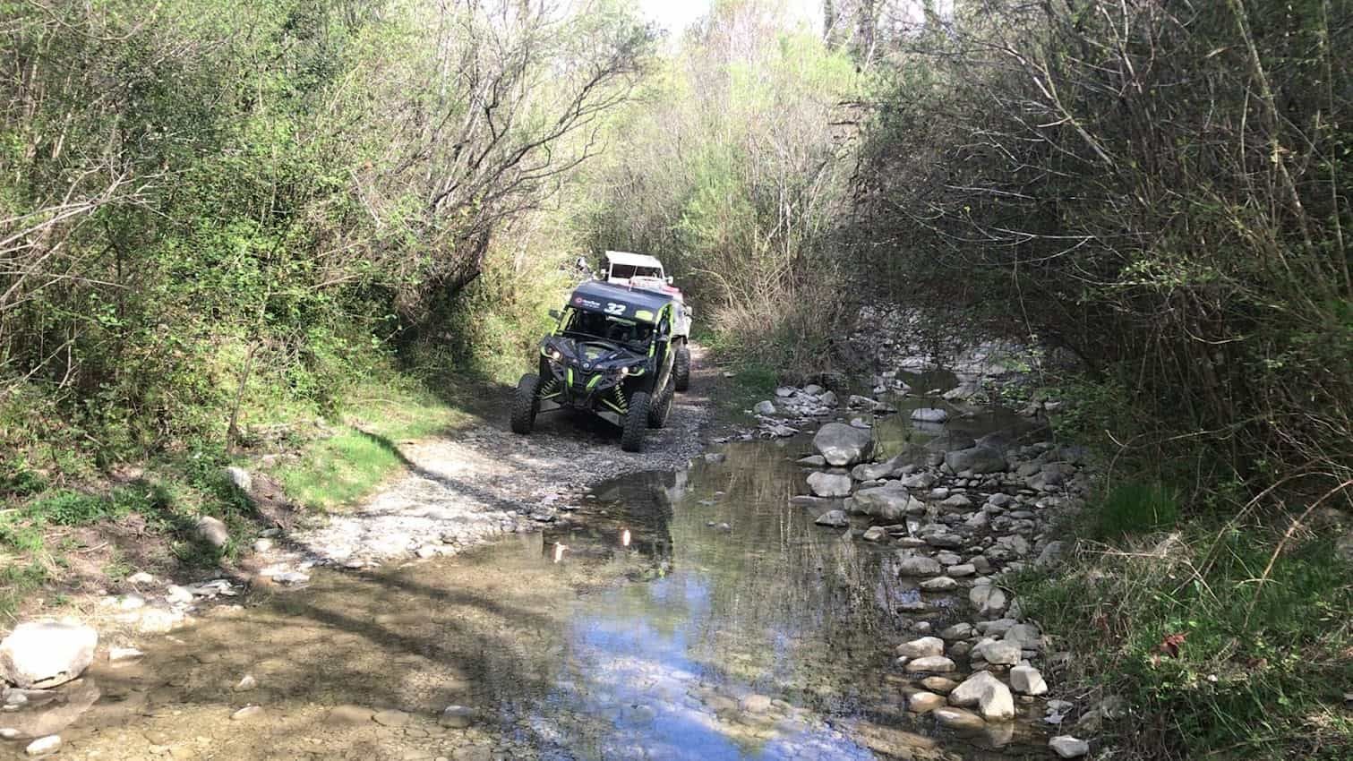 Dos vehículos todoterreno circulan a través de un arroyo poco profundo en una zona boscosa.