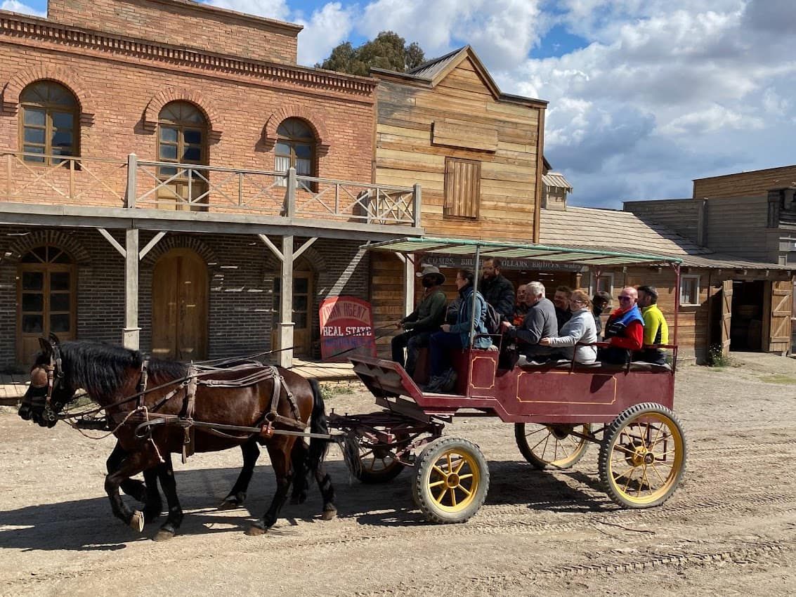 Carruaje tirado por caballos con pasajeros en un entorno urbano del oeste. Se ven edificios de ladrillo y madera.