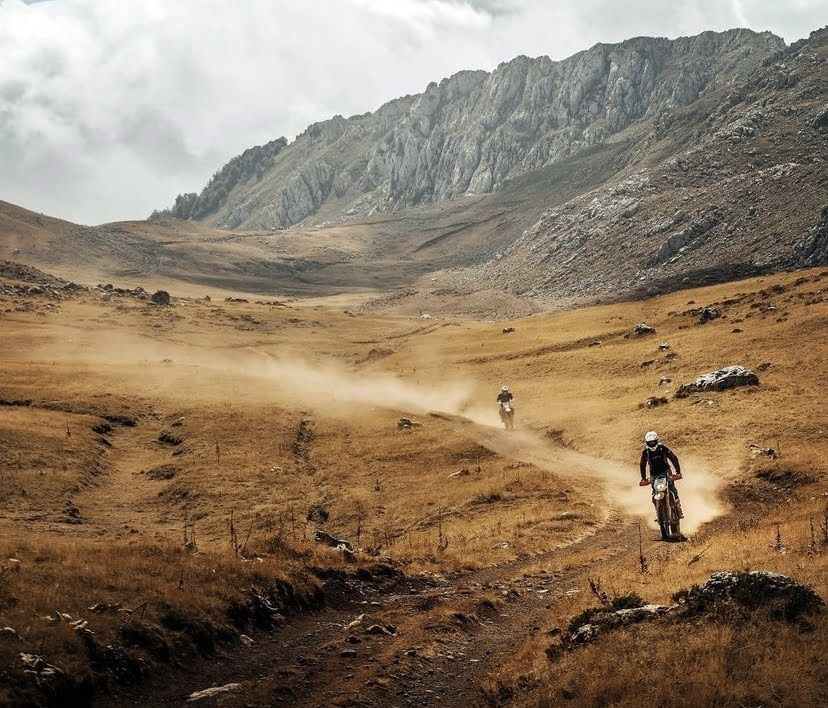 Dos motociclistas circulan por un camino de tierra en un paisaje montañoso, marrón y seco, levantando polvo.