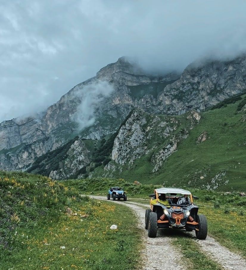 Dos vehículos todoterreno circulan por un sendero de montaña. Colinas verdes conducen a una montaña nublada.