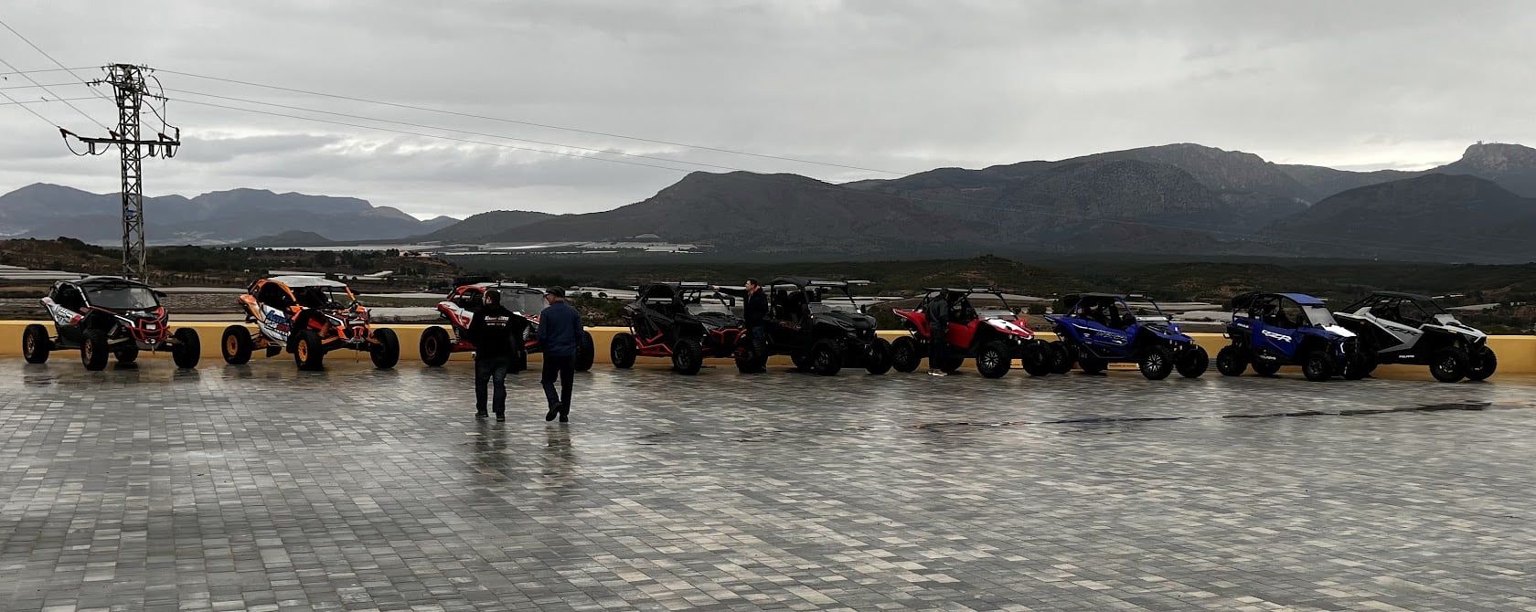 Una fila de vehículos todoterreno estacionados sobre una superficie mojada con dos personas caminando. Cordillera al fondo.