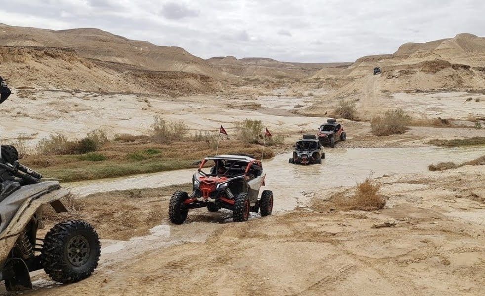 Cuatro vehículos todoterreno atraviesan el lecho fangoso de un río en un paisaje desértico.