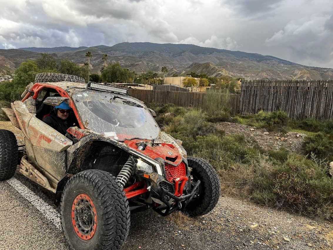Vehículo todoterreno cubierto de barro con conductor en un camino de tierra, montañas y una valla de madera al fondo.