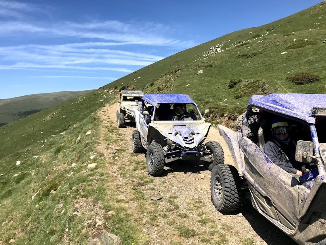 Vehículos todoterreno circulando por un camino de tierra a lo largo de una ladera cubierta de hierba bajo un cielo azul.