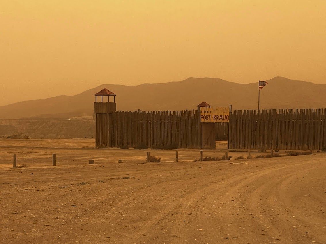 Fuerte de madera en un paisaje árido y polvoriento, con montañas al fondo bajo un cielo anaranjado.