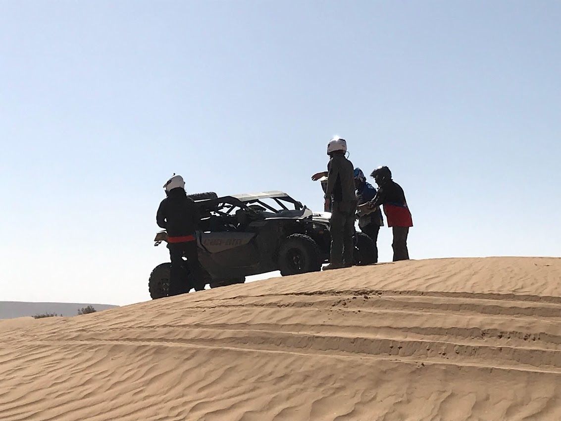 La gente se reúne alrededor de un vehículo todoterreno en una duna de arena bajo un cielo azul.
