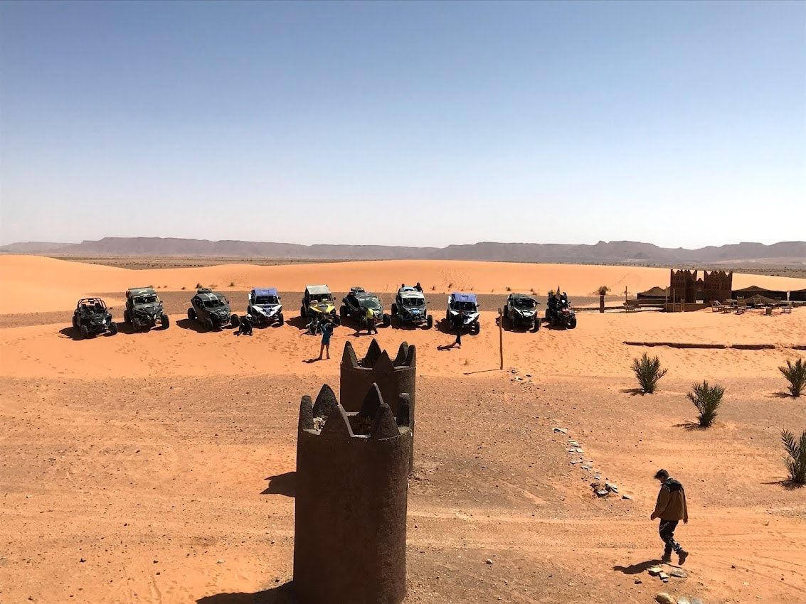 Vehículos todoterreno estacionados en un paisaje desértico con gente cerca y una estructura antigua en primer plano.