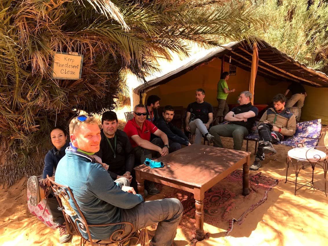 Grupo de personas relajándose en una zona sombreada con una tienda de campaña y una mesita. Palmeras al fondo.