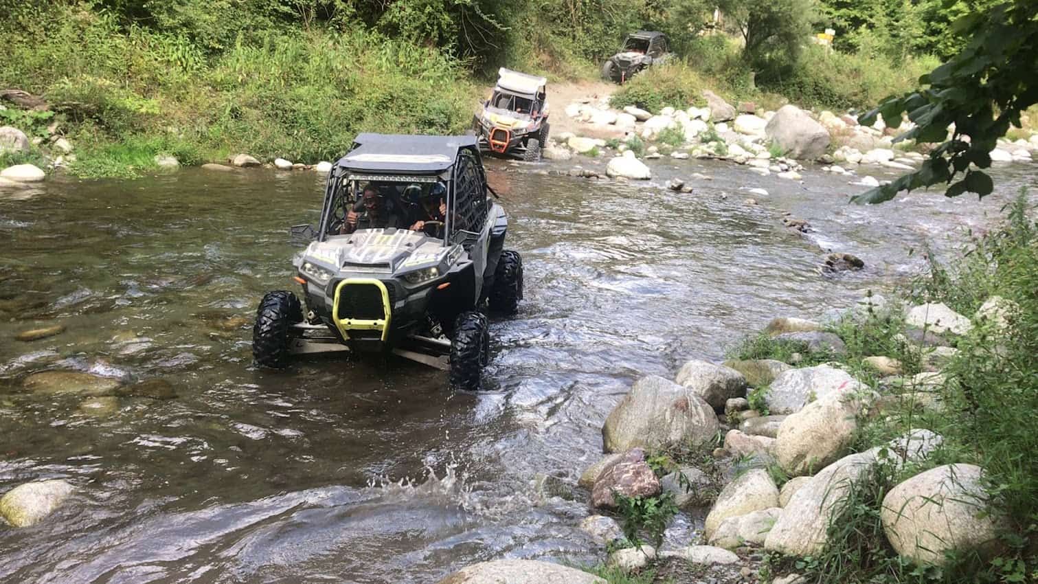 Vehículos todoterreno circulando a través de un río poco profundo, rodeado de árboles y rocas.
