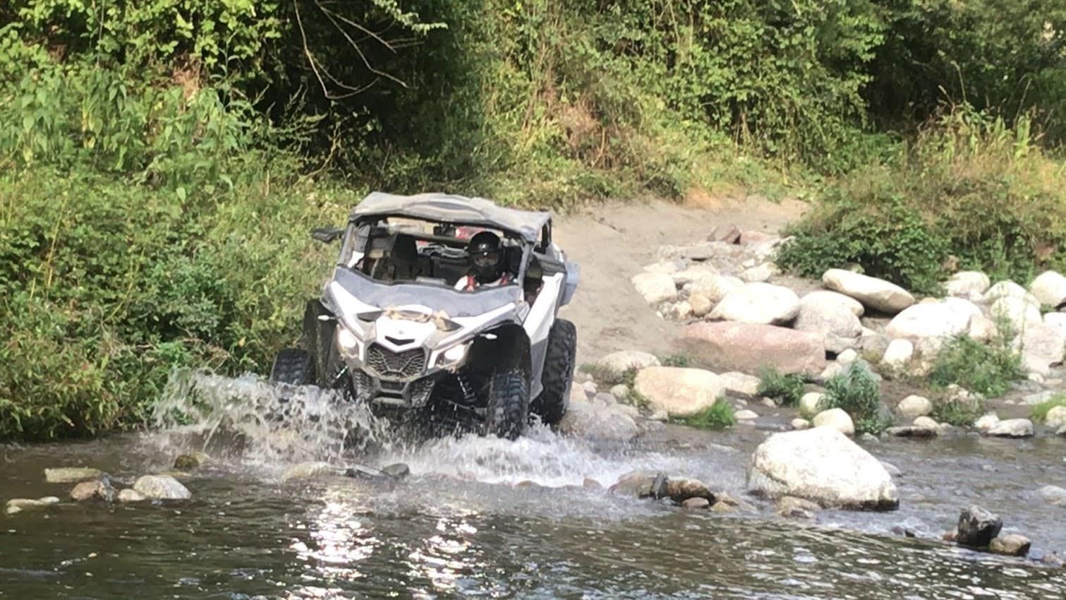 Vehículo todoterreno blanco vadeando un río poco profundo y rocoso con salpicaduras de agua, rodeado de vegetación verde.