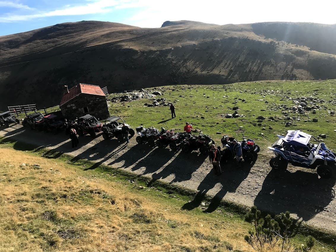 Vehículos todoterreno estacionados cerca de un edificio de piedra en una ladera cubierta de hierba, con montañas al fondo.
