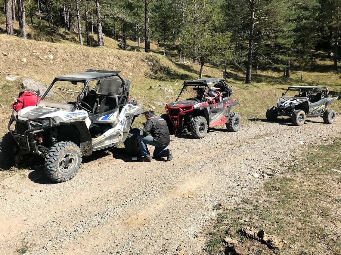 Tres cuatrimotos estacionadas en un camino de tierra en un bosque; personas trabajando en una de ellas.