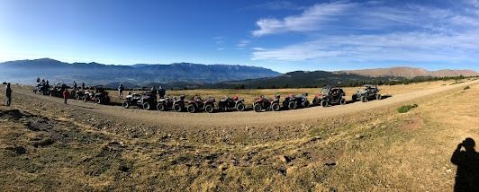 Un gran grupo de vehículos en un camino de tierra en un campo, con montañas al fondo bajo un cielo azul.