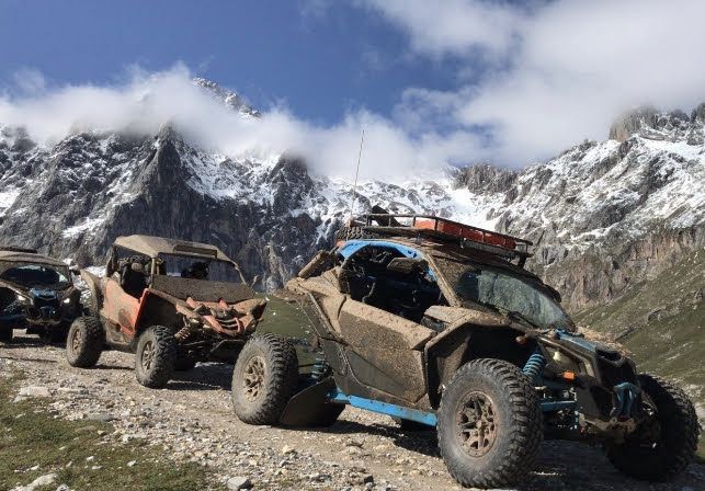 Vehículos todoterreno recorren un camino de tierra frente a montañas nevadas bajo un cielo azul.
