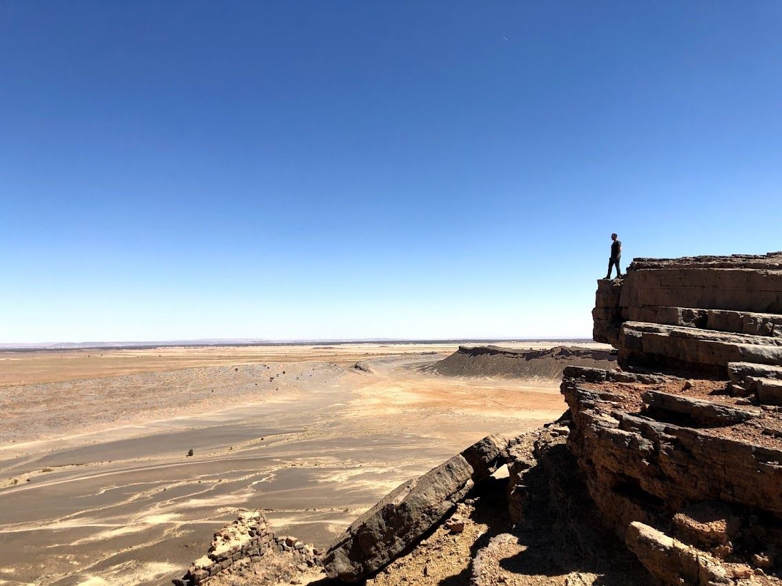 Una persona se encuentra de pie en un acantilado, contemplando un vasto paisaje desértico bajo un cielo azul despejado.