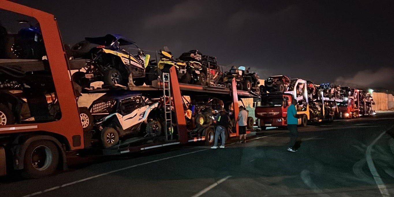 Varios camiones portacoches cargados con diversos vehículos por la noche. Hay gente de pie cerca de los camiones.