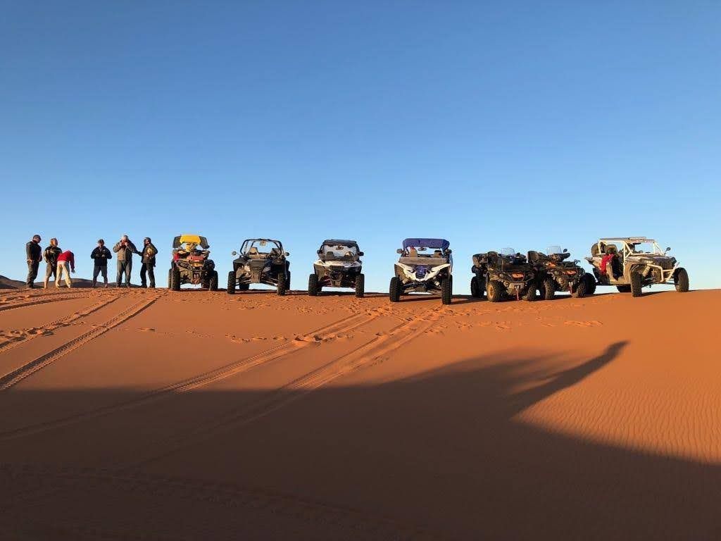 Grupo de personas y vehículos todoterreno sobre una duna de arena roja bajo un cielo azul.
