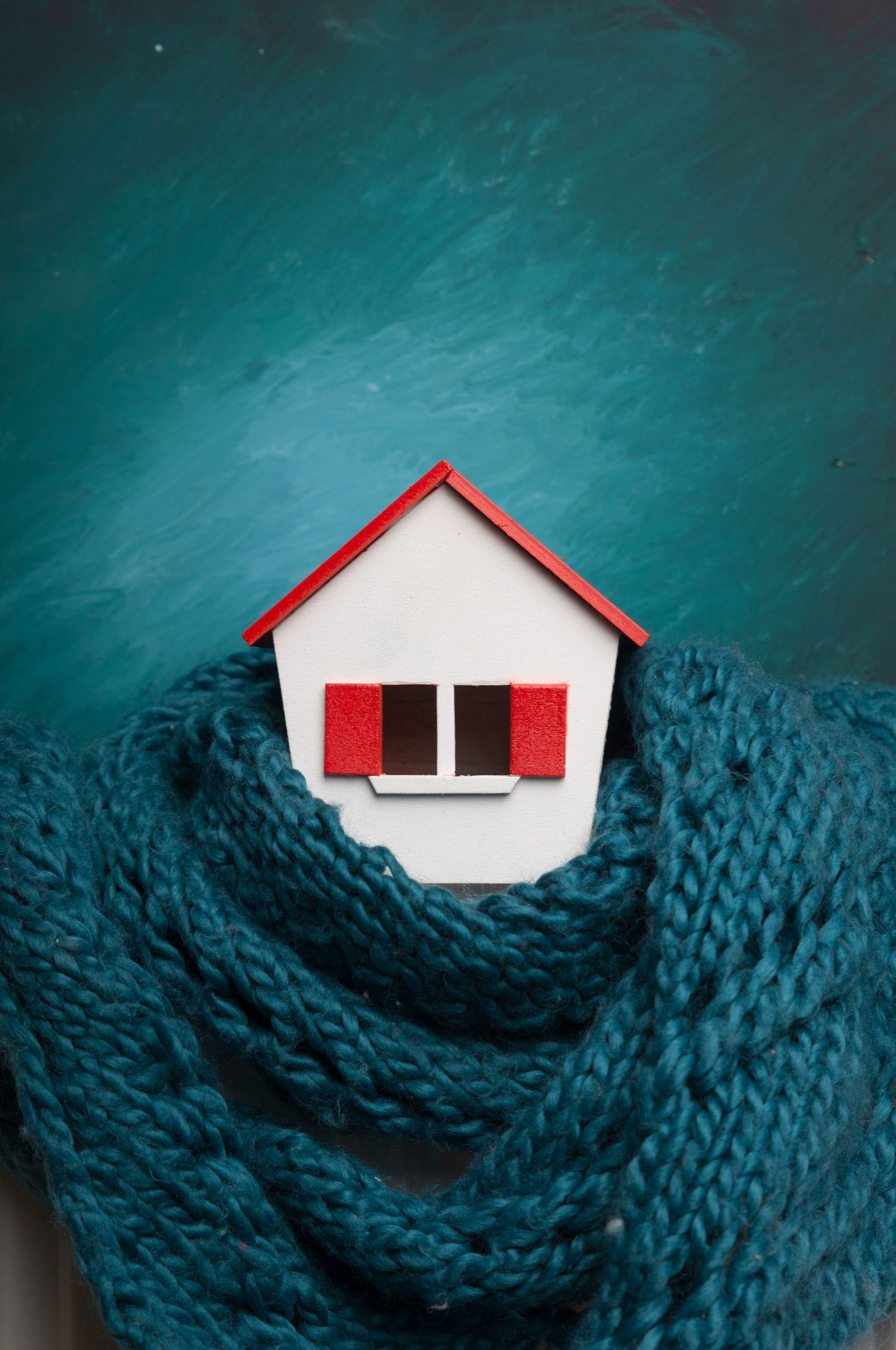 Maison blanche aux volets rouges, enveloppée d'une écharpe en tricot bleu sur fond turquoise.