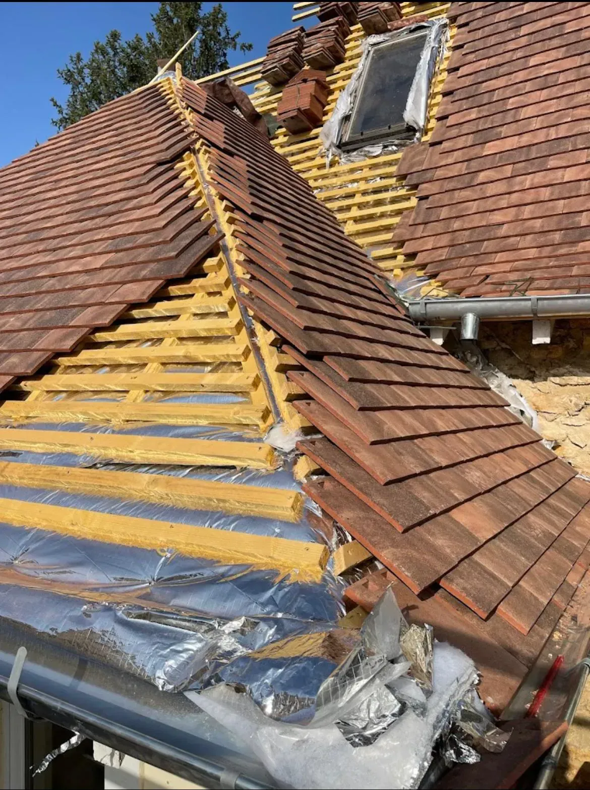 Toiture en construction : tuiles rouges, isolation apparente, poutres en bois, fenêtre de toit.