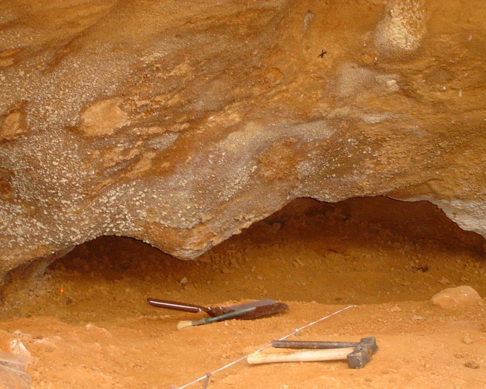 Un martillo y una pala están tirados en la tierra frente a una cueva.