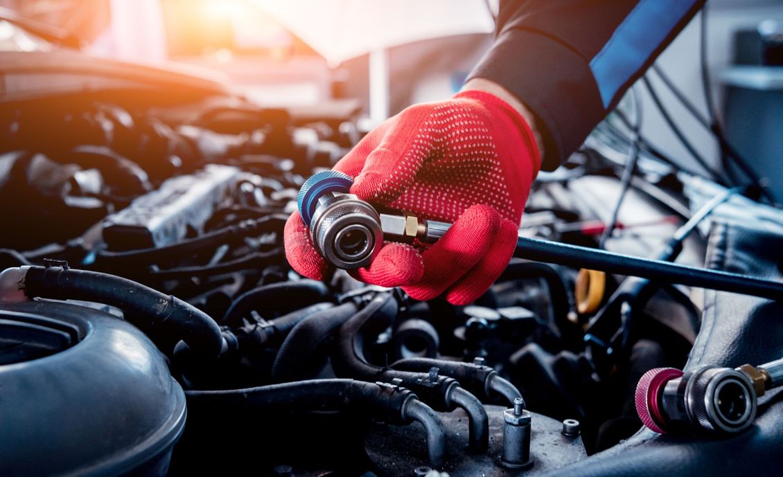 Main gantée tenant un tuyau de recharge de climatisation au-dessus d'un moteur de voiture.