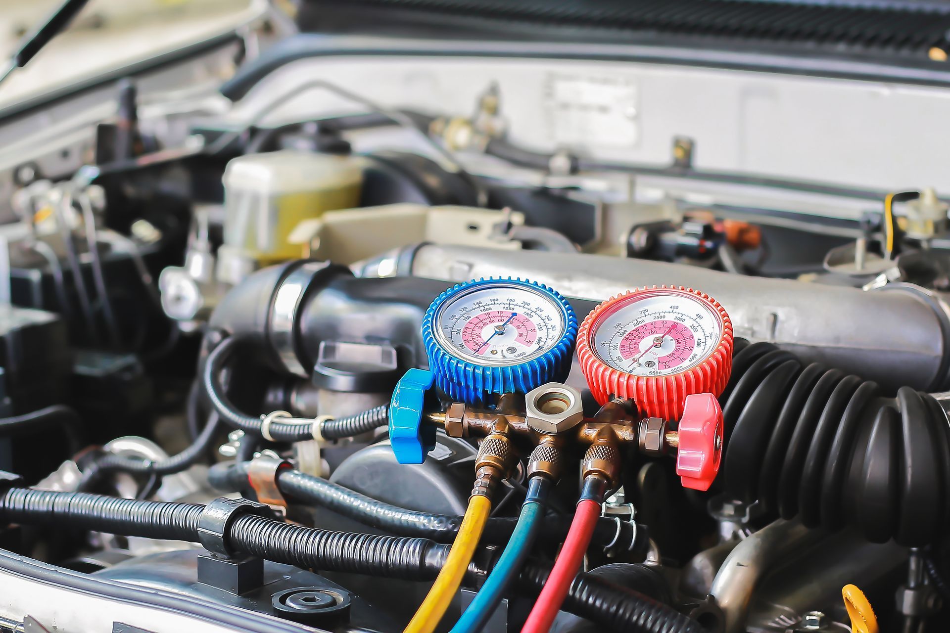 Moteur de voiture avec manomètres à collecteur ouvert, cadrans rouge et bleu reliés par des tuyaux pour l'entretien de la climatisation.