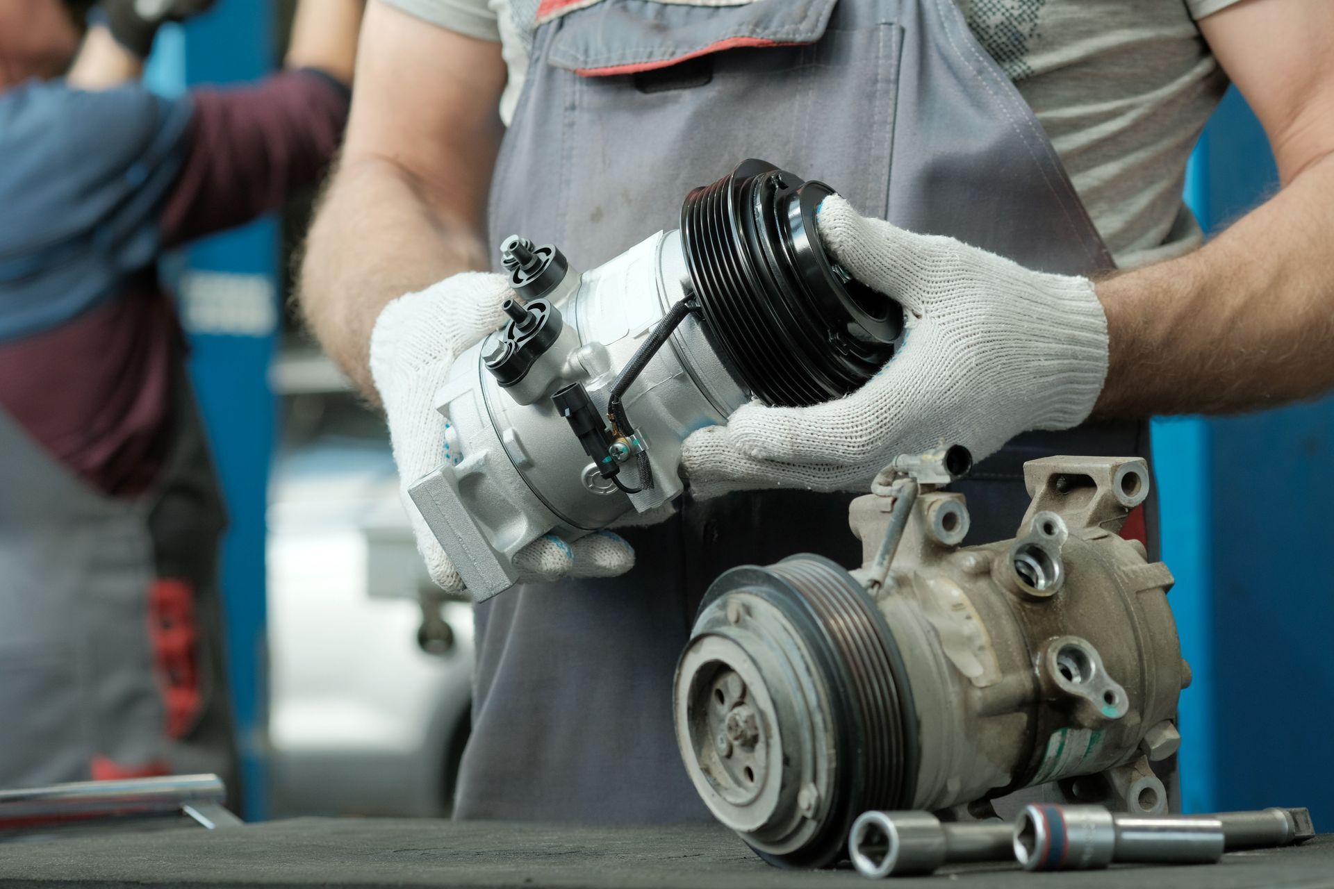 Un mécanicien portant des gants gris manipule un moteur de climatisation de voiture sur un établi, entouré d'outils.